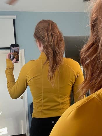 A person with long, wavy hair is taking a mirror selfie from the back, showing off their yellow long-sleeve athletic top