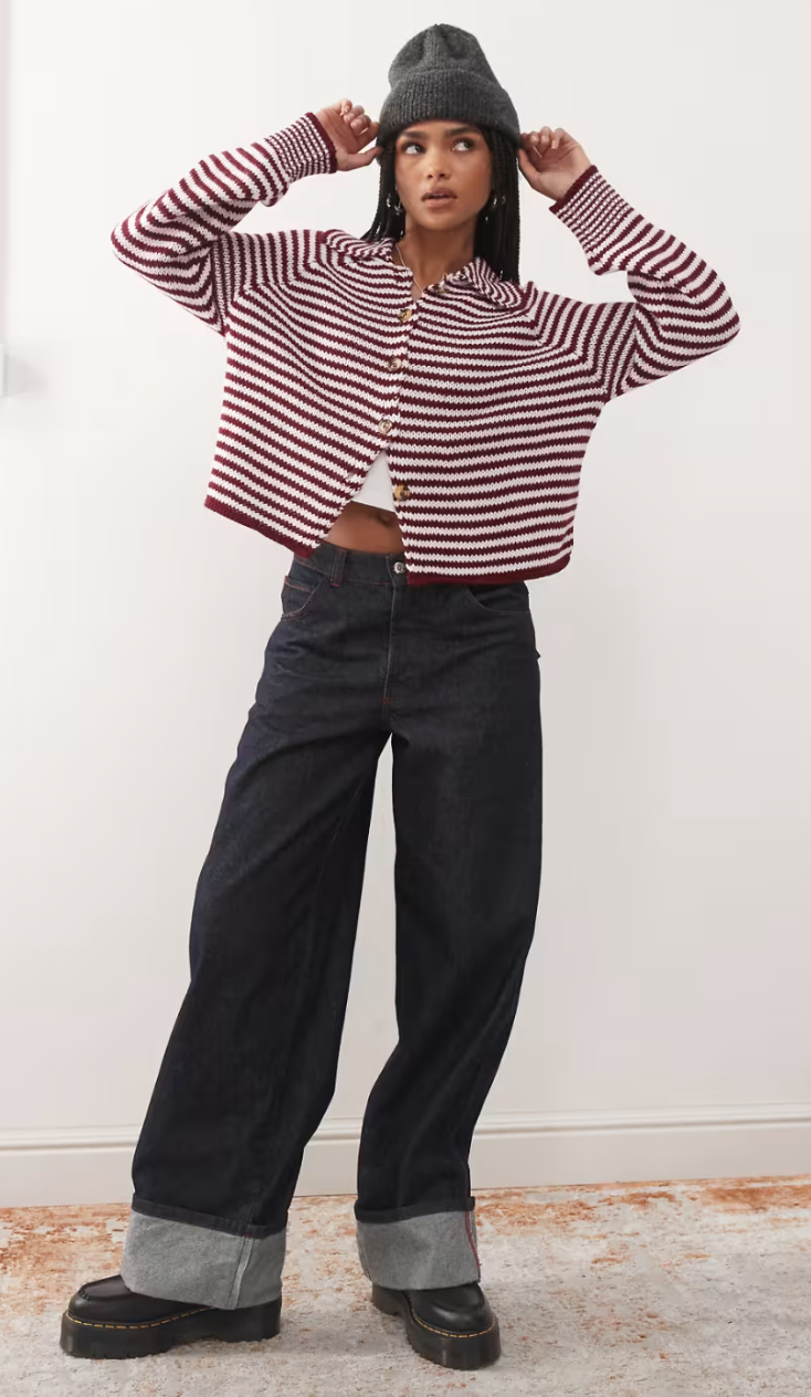 Model wearing a striped button-up sweater, cuffed wide-leg jeans, a white crop top, platform boots, and a beanie, posing indoors