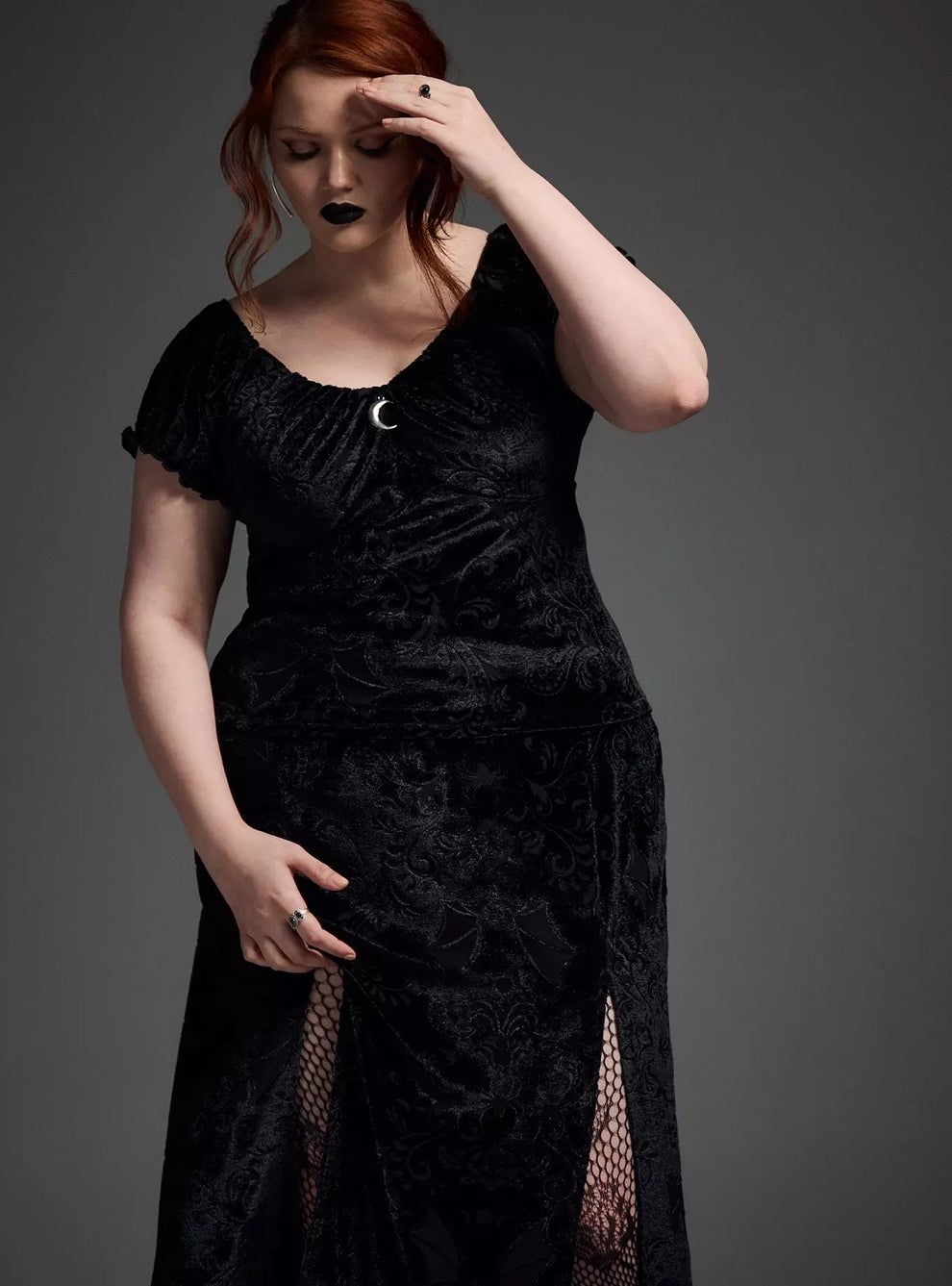 Person wearing a black, patterned dress with fishnet detail, standing in a contemplative pose