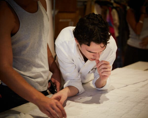 Mr. Ives, in a white shirt with sleeves rolled up, leans over a workroom table studying a garment in progress. A studio worker stands next to him, scissors in hand.