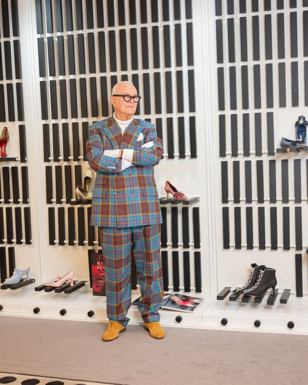 Mr. Blahnik in a blue, red and yellow plaid suit stands, his arms crossed, in front of wall display of assorted shoes. 