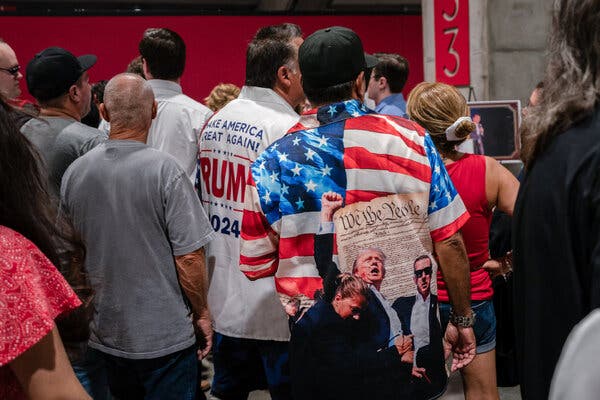 A group of people are seen from behind at Charlie Kirk's memorial service. One is wearing a shirt with an American flag, the constitution and an image of President Trump raising his fist. Another man is wearing a shirt that says Make America Great Again Trump 2024.