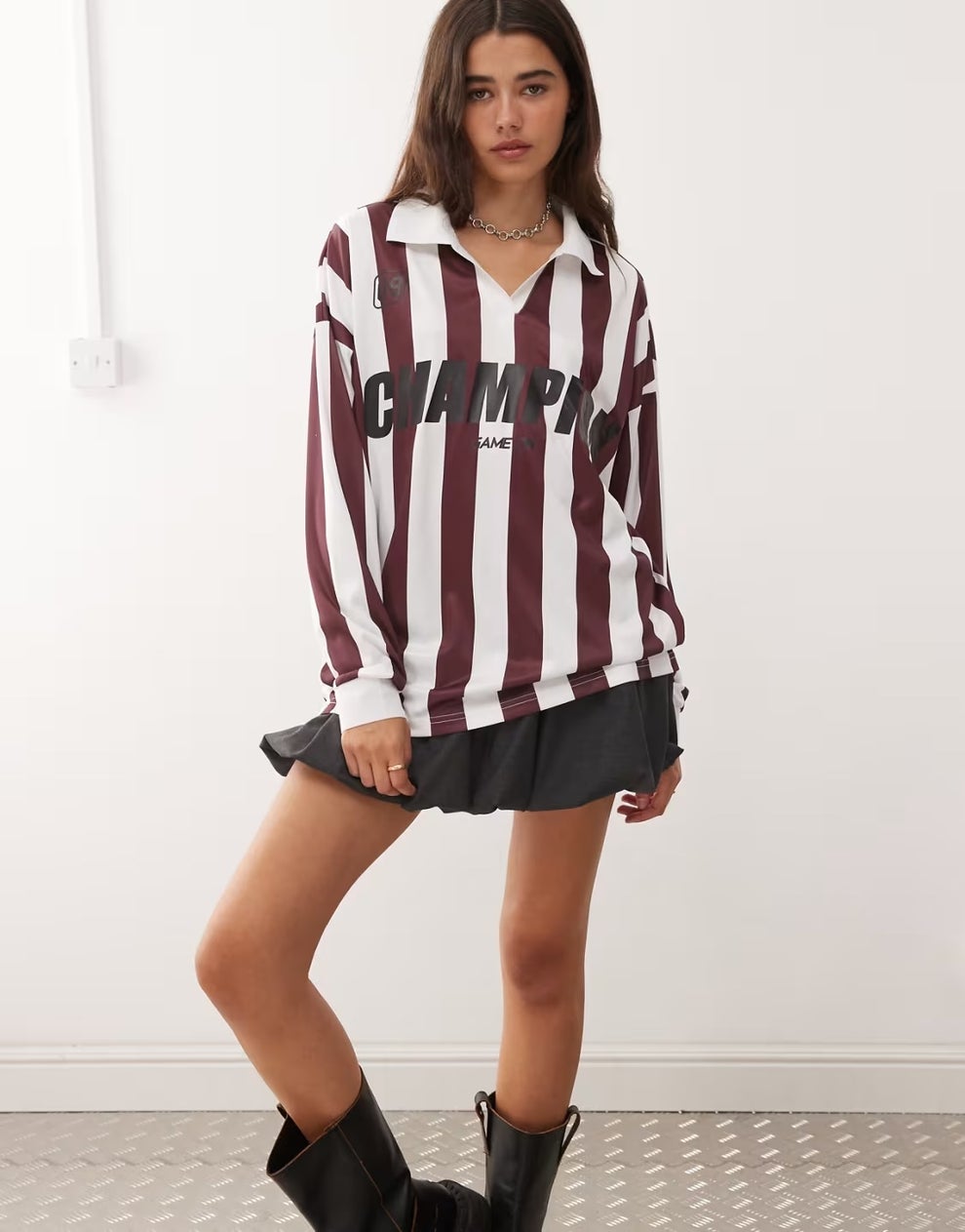 Person wearing a striped, long-sleeve top with "CHAMPION" text, a short pleated skirt, and black boots, standing in a casual pose