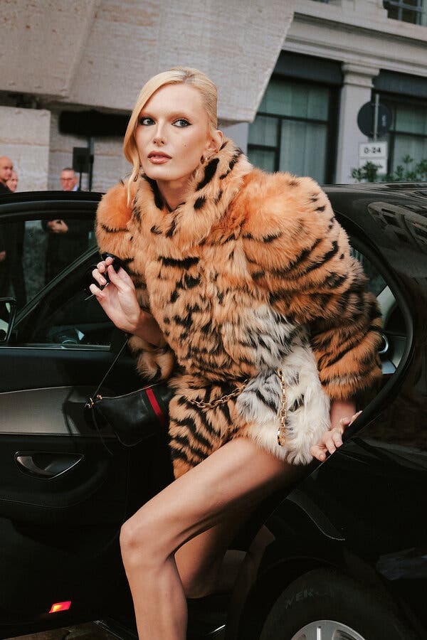 A blonde woman in a tiger-stripe fur-look jacket is climbing out of a dark automobile. 