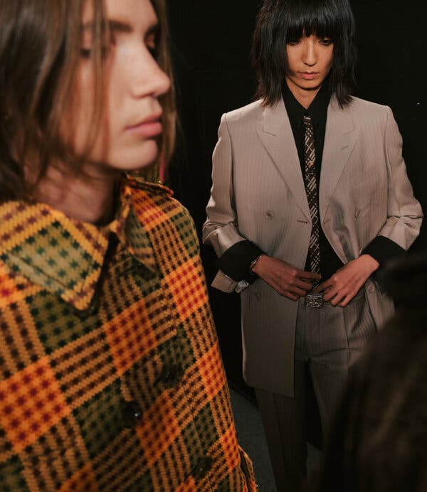 Two models backstage at a fashion show. One model, left, is wearing a plaid coat in autumnal colors. Another, right, is wearing a gray suit, a black shirt and a gray-and-black-plaid tie. 