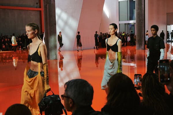 A line of models, led by two in black bras and droopy skirts hung by straps from their shoulders, walk a reflective orange-red runway.