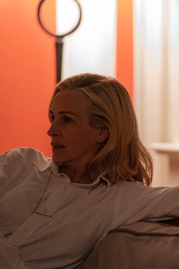 In a still from the film, Ms. Roberts’s character leans back on a sofa. She’s wearing a white button-up shirt with a long gold chain around her neck. 
