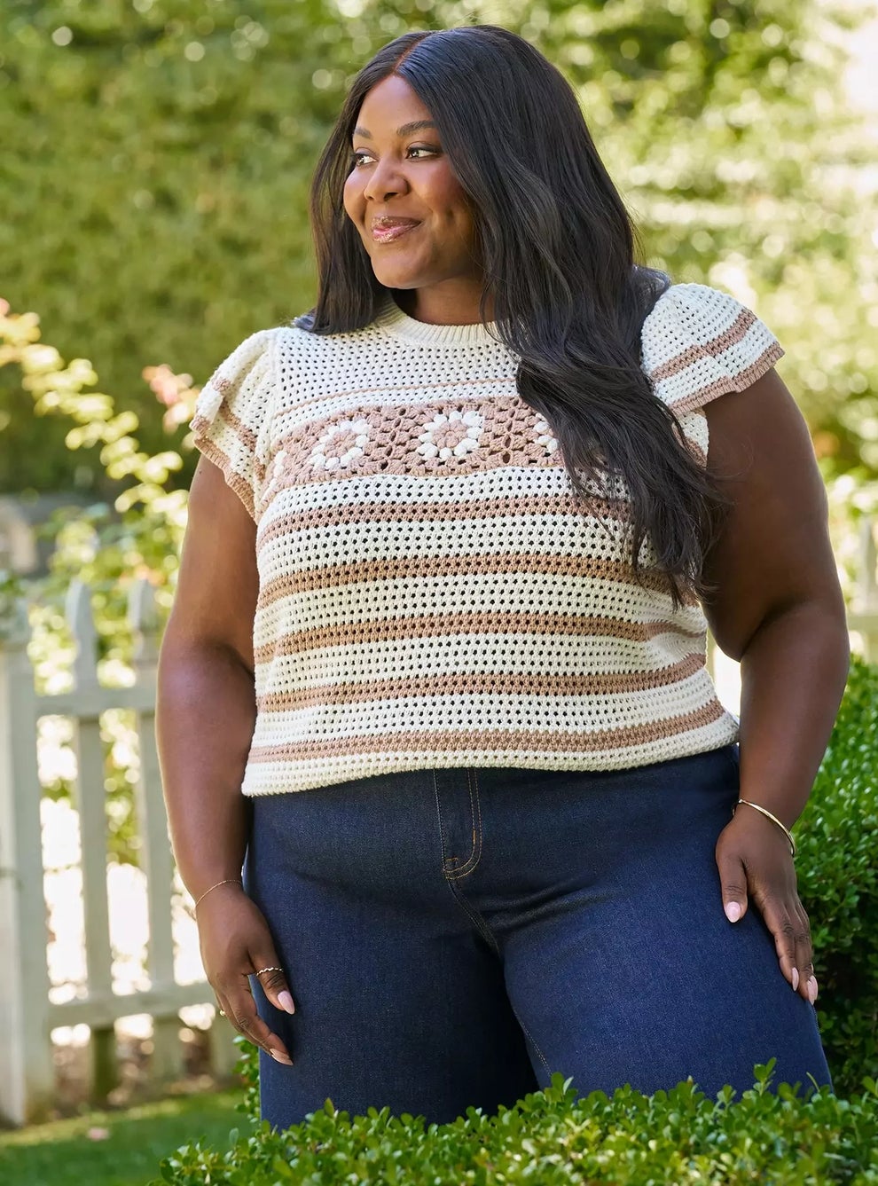 Person outdoors wearing a crochet floral top and jeans, standing in a garden setting, smiling towards the distance