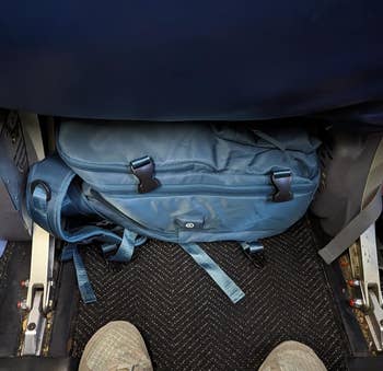 A reviewer showing how well their blue backpack fits underneath the airplane seat in front of them