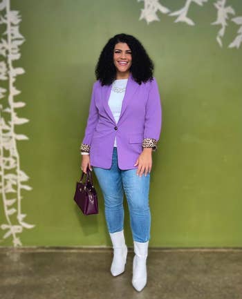 Person smiling in a chic purple blazer with leopard print cuffs, holding a handbag, wearing jeans and white ankle boots, against a leafy backdrop
