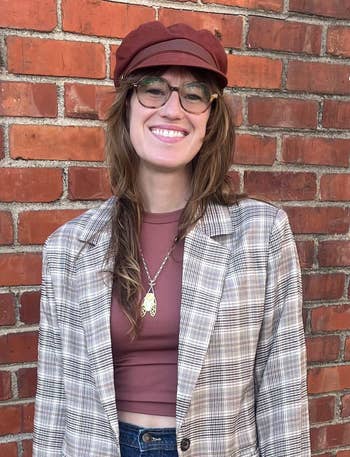 Smiling woman in glasses, wearing a plaid blazer, maroon hat, and a necklace, stands against a brick wall