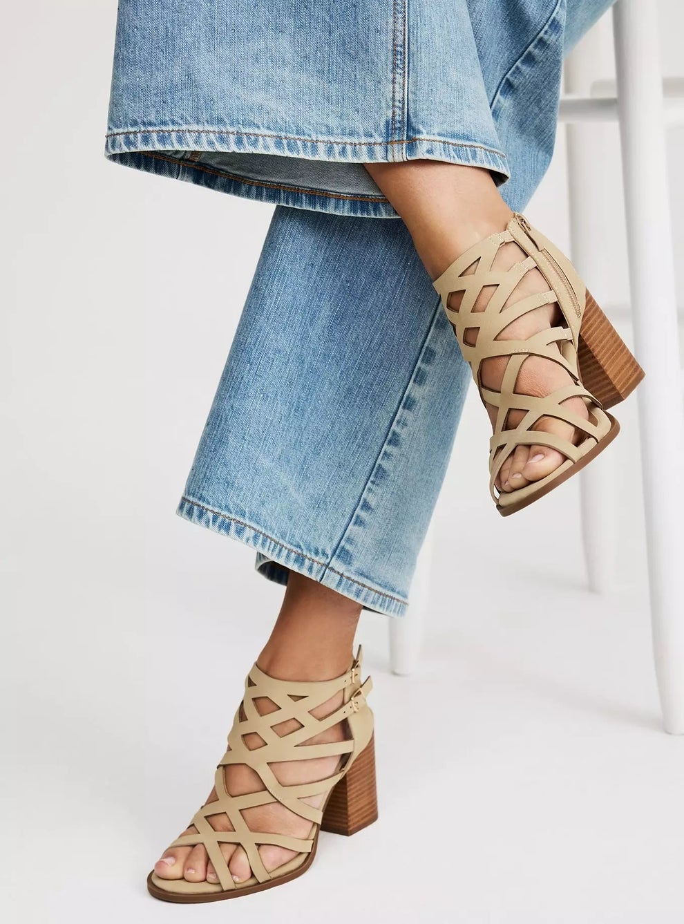 Model wearing tan strappy high-heeled sandals with wide-leg jeans, seated on a stool