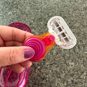 A hand holds a razor with a pink handle, featuring three blades. A matching pink razor cover is visible on the countertop