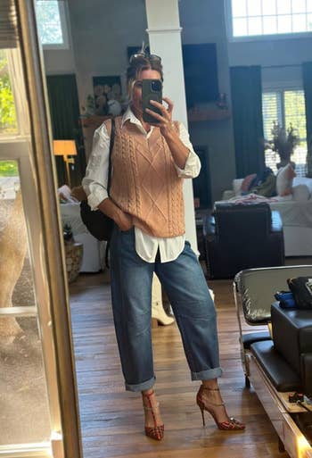 Person taking a mirror selfie in casual attire, including a cable-knit vest, white shirt, jeans, and high heels, standing in a stylish living room