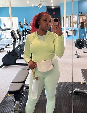Person in a gym wearing athletic wear, holding a water bottle and standing in front of exercise equipment