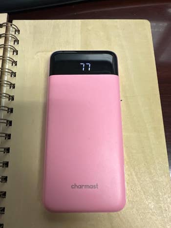 Portable power bank on a notebook, digital display shows 77 percent charge, brand name visible. Ideal for tech shopping features