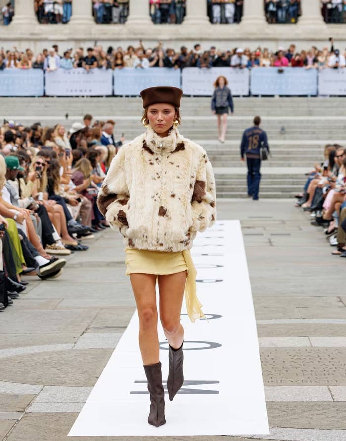 Model walks runway in faux fur coat, short skirt, and boots.