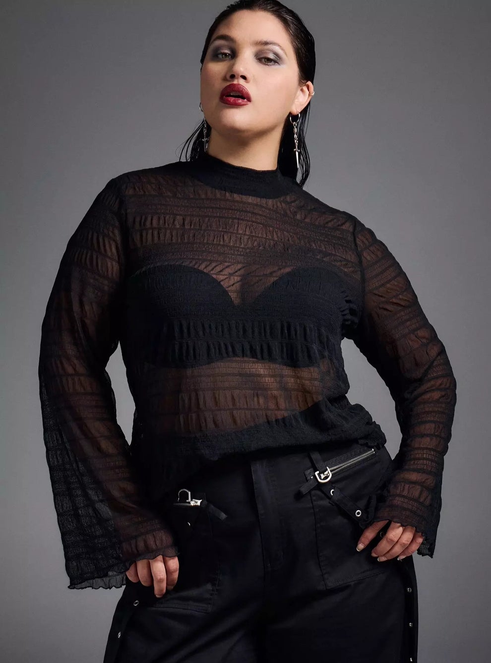 Person in a sheer, long-sleeve, black top with layered detailing and black pants, posing confidently against a plain background