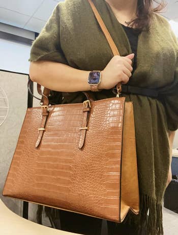 A person holds a large brown crocodile-patterned handbag 