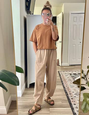 Person wearing casual oversized top and loose pants, with sandals, taking a mirror selfie in a cozy room