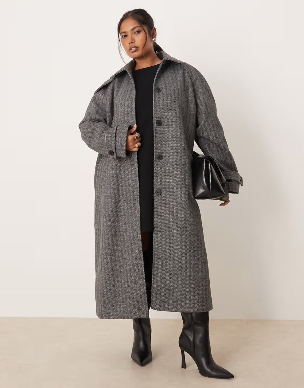 Person models long trench coat, black dress, and knee-high boots, holding a shiny handbag.