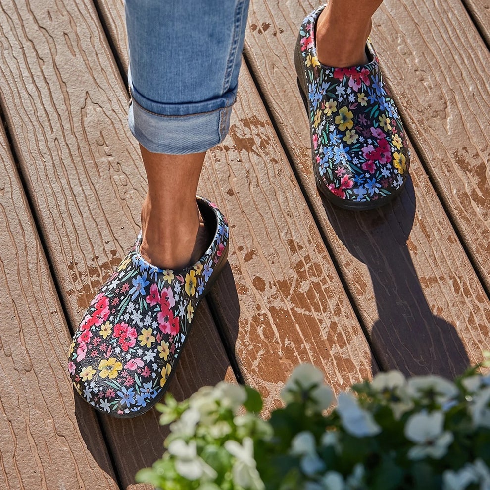Person wearing floral pattern clogs and rolled-up jeans stands on wooden decking. White flowers visible in the foreground