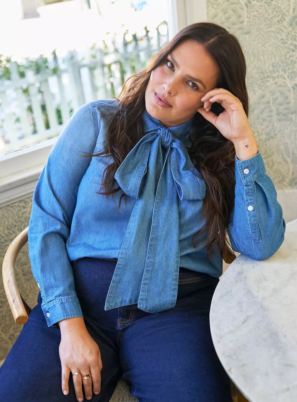 Person seated at a table wearing a denim shirt with a large bow, looking directly at the camera with a relaxed expression