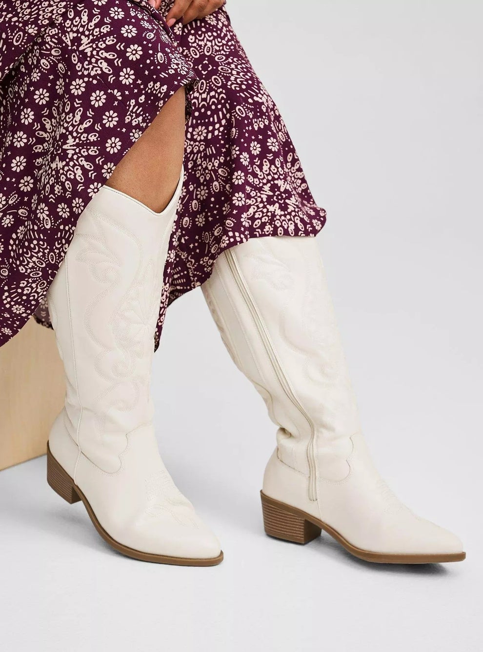 Person sitting, wearing knee-high cowboy boots with a floral-patterned skirt. The boots have a detailed embroidered design