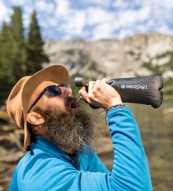 Person outdoors using a LifeStraw water filter bottle by a lake, wearing a hat and jacket, enjoying nature