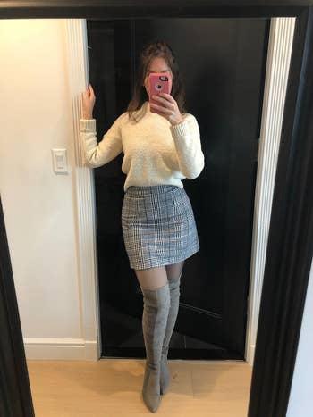 reviewer taking a mirror selfie, wearing a white sweater, plaid skirt, and thigh-high suede boots