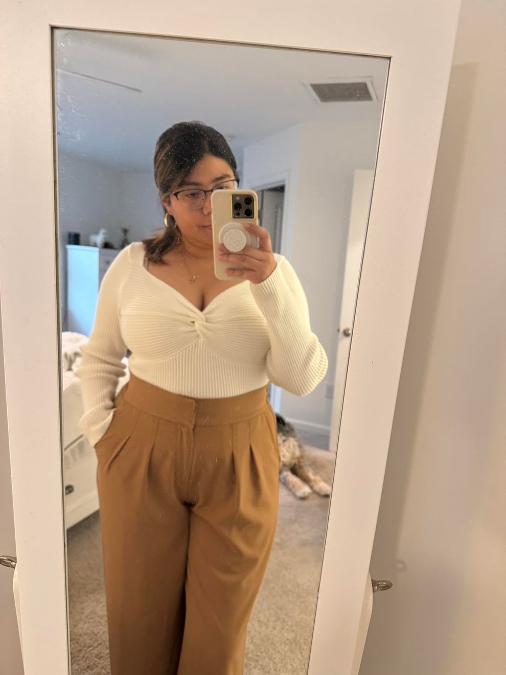 Person taking a mirror selfie in a white sweater and tan pants, standing in a bedroom