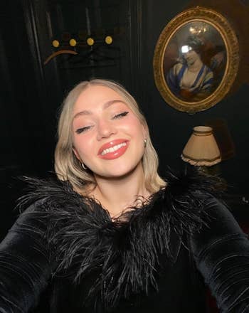 Person smiling in a room, wearing a black top with feather details. Vintage decor in the background. Used in Shopping article