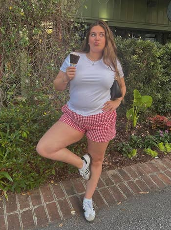 Person outdoors holding an ice cream cone, wearing a white t-shirt, red gingham shorts, and sneakers, striking a playful pose