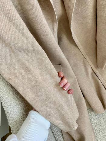 Close-up of a person holding a tan textured fabric coat. The person is wearing a white ribbed sleeve