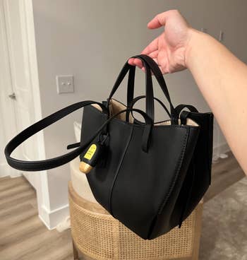 Hand holding a stylish black handbag with a yellow tag, set in a modern room, highlighting its sleek design