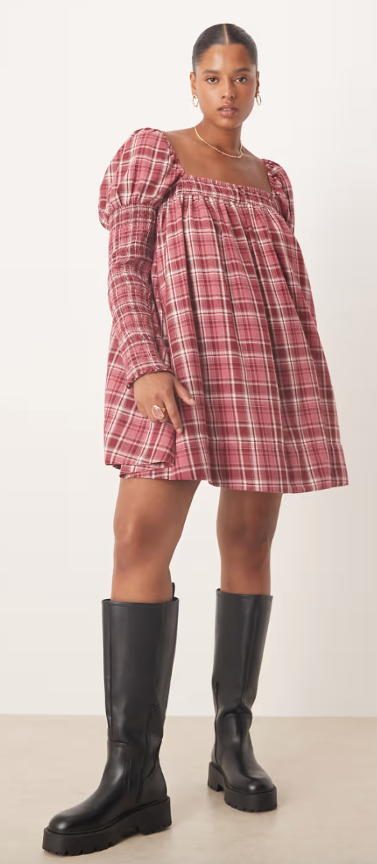 Person in a plaid, puff-sleeve dress and tall black boots, striking a casual pose against a plain background