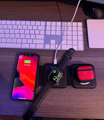 iPhone displaying battery percentage, Apple Watch on a charging pad, and AirPods in a case on a wooden desk next to a keyboard