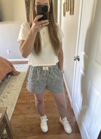 Person taking a mirror selfie wearing a white crop top, checkered shorts, and sneakers, standing in a bedroom setting