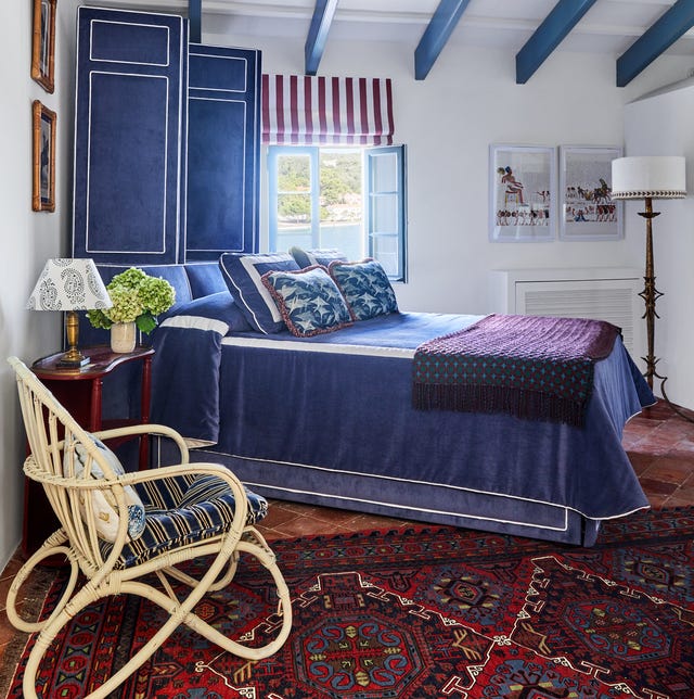 white room with blue ceiling beams and a bed set on an angle with a lush royal blue velvet cover with white trim and purple throw at the foot and matching blue folding screen with white trim behind the bed and a terracotta floor w ith a majestic blue and red patterned rug and a simple rattan chair in the foreground