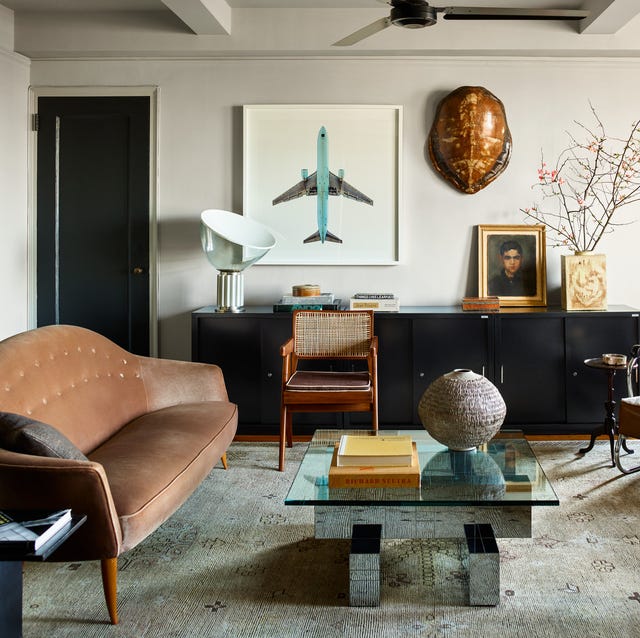 a living room with a long black console on the back wall with art and book around it and a worn leather sofa on the left and a low geometric cocktail table with a glass top in fron on a faded rug and old metal and leather armchair at right