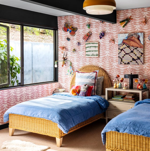 twin rattan beds with oval headboard and blue coverlets against a pink patterned wallpaper and a nightstand in between and a long window and bug artwork and objects on the wall
