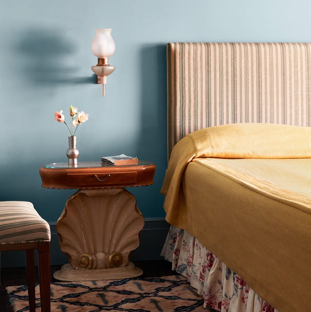 bedroom with blue wall and a bed with tall striped headboard and flowered bed skirt and plain white coverlet and a patterned pink and blue carpet and a nightstand with an oval wood top and a seashell pedestal