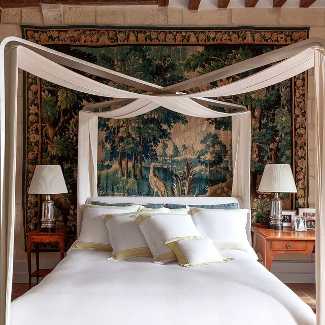 a white linen bed with a ribbon canopy draped in an x overhead and a large tapestry on the wall behind and two small mismatched sidetables and silver base lamps