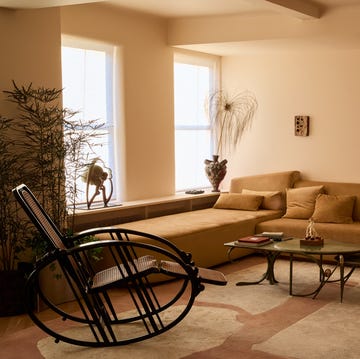 living room with long tan colored sofa built along cream colored walls with two windows and a rocking chair