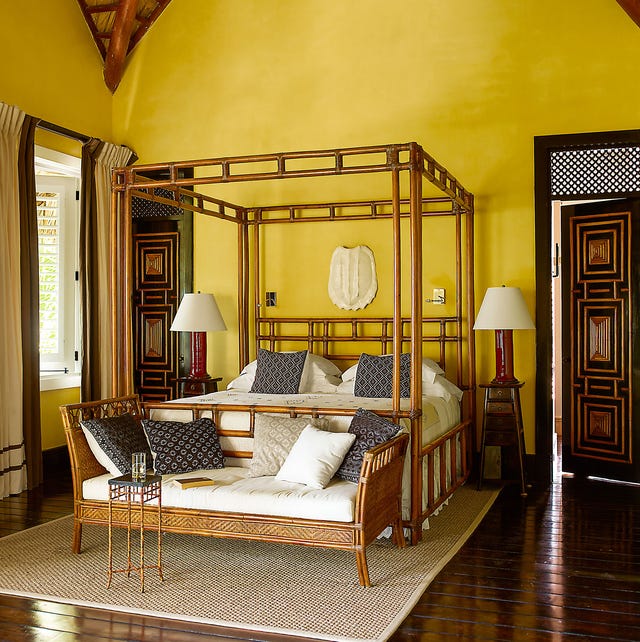 yellow painted room with bamboo cane four poster bed and matching empire looking settee at the foot and dark wood floors and carved black doors with wood insets