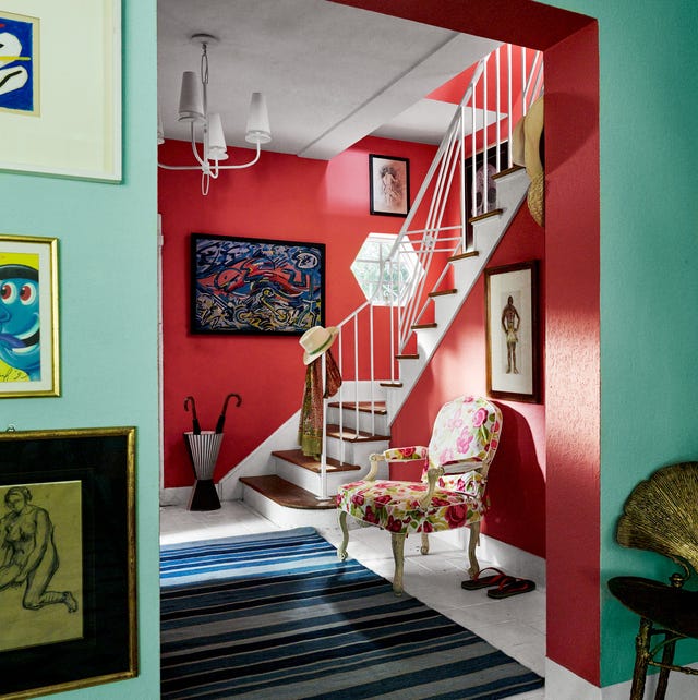 view from a green painted room with artwork into a red painted entry with stairs going up and a floral fauteuil next to it and a striped rug in blue hues and artwork on the walls and a simple chandelier