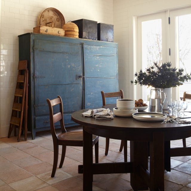 a round wood table with plates and glasses on it and a rustic blue painted chest behind it with old fashioned boxes and bins and french doors to the back right