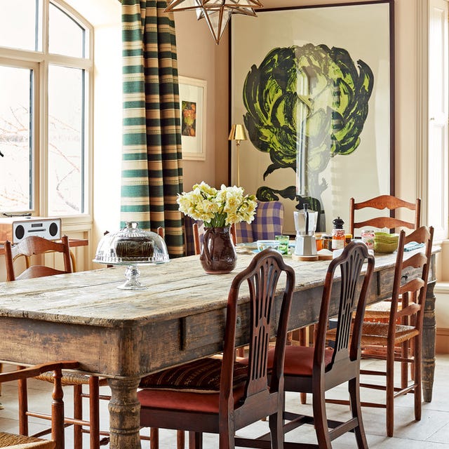 a rustic dining room table with chairs and a green and cream striped curtains at the window and a large painting of an artichoke in a vivid green at the back and a star chandelier overhead
