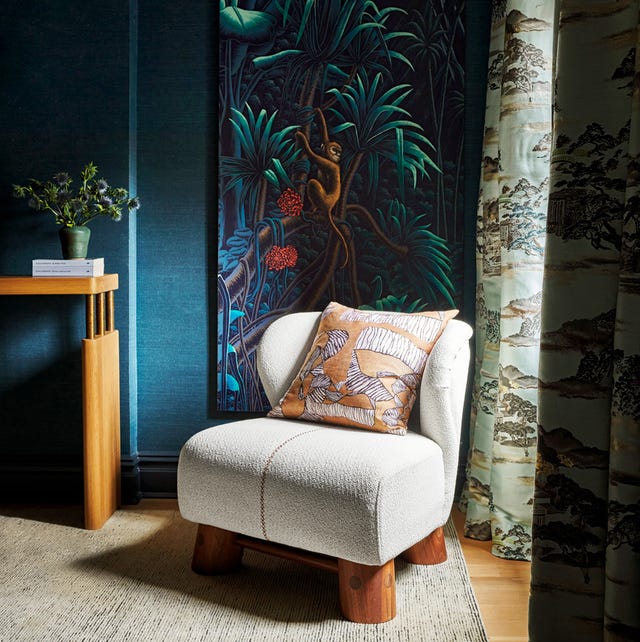 white upholstered chair with thick wood beam legs on a light striated carpet agsinst a blue textured wall with a monkey artrwork and toile curtains in a moody corner