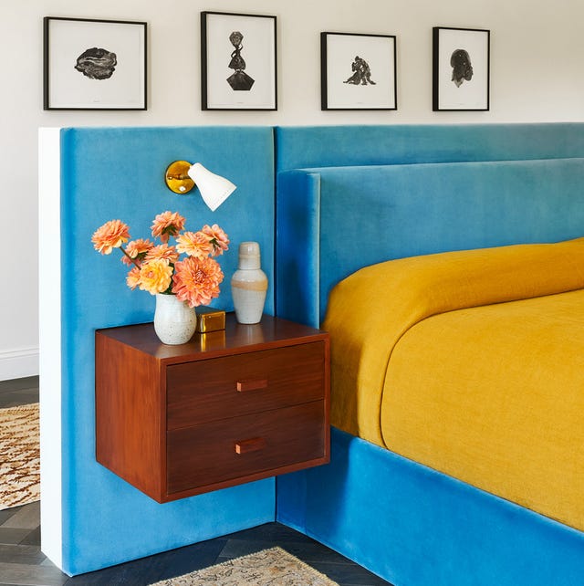 upholstered platform bed in a blue velvet with a yellow cover and a floating mounted two drawer night stand with a small white vase with flowers on it and a small mounted white spotlight
